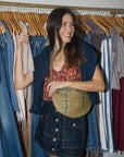 A woman stands in front of a clothing rack wearing a navy sleeveless blazer, a red floral top, a dark denim skirt, and a green woven crossbody bag.