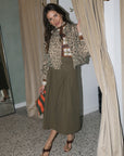 A woman posing in a fitting room wearing a leopard-print cropped jacket, a brown and white striped tie, an olive green midi skirt, and brown strappy sandals, holding a striped clutch.
