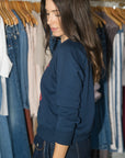 A woman with long dark brown hair is shown from the side, wearing a navy blue crewneck sweatshirt and dark wash jeans, standing in front of a clothing rack filled with jeans and other garments.