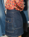 A close-up of a person wearing a dark blue denim mini skirt with a front flap pocket and contrast stitching, paired with a red floral blouse. The background shows a clothing rack with blue denim garments.