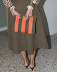 A person wearing a leopard-print top and olive green midi skirt holds a striped clutch bag with vertical orange and dark green stripes. The person is wearing brown heeled sandals on a speckled terrazzo floor.