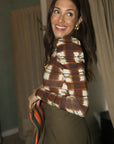 A woman wearing a brown, beige, and white patterned long-sleeve top and olive green skirt, holding a striped clutch bag in an indoor setting.