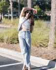 Woman in light blue jeans and a white top standing on a street with palm trees in the background, full body front