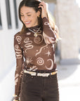 Woman wearing a brown patterned long-sleeve top, Cape Necklace White and dark jeans outdoors.