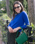Woman in a blue sweater and sunglasses holding a green handbag outdoors.