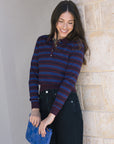 Woman wearing a striped sweater and holding a blue clutch against a stone wall.