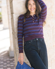 Woman wearing a striped sweater and jeans, holding a blue clutch against a stone wall.