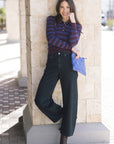 Woman in a striped shirt and black jeans leaning against a stone wall.