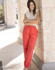 Woman wearing red pants and a beige top standing against a stone wall.