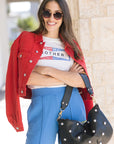 Woman wearing a red jacket, white graphic t-shirt, blue pants, and sunglasses, holding a black studded handbag.