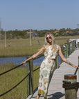 Woman in a patterned set standing on a wooden dock by a body of water with grass and trees in the background.