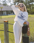 Woman in a white sweatshirt with a logo and white pants standing outdoors near a wooden post.