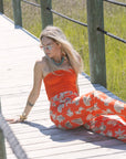 Woman in Tracey Necklace and an orange top and pants with a pattern sitting on a wooden bridge.