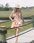 Woman in a patterned dress and hat standing on a wooden dock by a body of water. full body view