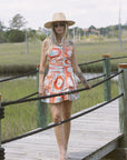 Woman in a patterned dress and hat standing on a wooden bridge over a waterway.