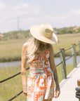 Woman in a patterned dress and sun hat standing on a wooden boardwalk with a scenic background. Close up front