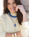 Woman wearing a white sweater, blue beaded necklace, and gold bracelets with a neutral background