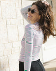 Woman wearing a patterned long-sleeve top and sunglasses, standing against a light stone wall.