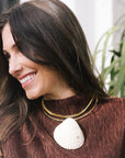 A woman with long dark brown hair smiling, wearinga large white seashell pendant necklace and a smaller gold chain.