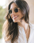 A smiling woman with long brown hair wearing round sunglasses with a clear frame and dark lenses, paired with a white striped shirt and a gold necklace.