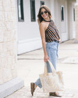 A woman wearing a brown and black striped top, light blue jeans, and brown boots is walking outdoors while holding a cream and gold tote bag.