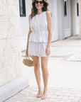 A woman stands outdoors on a paved walkway wearing a sleeveless vertically striped set. She is holding a small tan handbag and wearing tan high-heeled sandals.