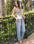 A woman posing outdoors in a brown and white striped crop top, light blue wide-leg jeans, and brown slide sandals, carrying a silver fanny pack.