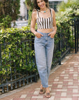 A woman stands on a paved walkway outdoors, wearing a brown and white vertically striped crop top with thin shoulder straps, light-wash blue jeans, and brown heeled sandals.