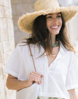 A woman wearing a wide-brimmed straw hat, a white short-sleeve button-up shirt, and a green patterned skirt, holding a white shoulder bag against a light stone wall background.