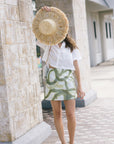 A woman stands outdoors on a paved walkway beside a stone column, holding a large straw hat over her face. She is wearing a white short-sleeve top and a white mini skirt with an abstract green pattern.