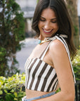 A woman wearing a brown and white vertically striped halter crop top with tie straps, paired with light blue jeans, photographed outdoors in front of a blurred garden background. side view