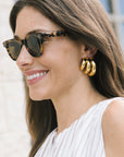 A close-up side profile of a woman wearing gold triple-hoop earrings and tortoiseshell sunglasses, with a white pleated top against a softly blurred light background.