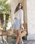 A woman stands on a stone patio wearing a cream long-sleeve button-up shirt, a light blue flared mini skirt, sunglasses, and blue slide sandals, while holding a tan handbag.