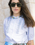 Woman wearing a light blue striped blouse with decorative elements, standing against a stone wall.