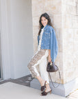 Woman in denim jacket and white outfit standing against a stone wall.