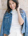 Woman wearing a denim jacket over a white top against a stone wall.