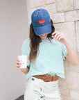 Woman wearing a blue cap, green striped shirt, and white skirt with floral pattern, holding a coffee cup.