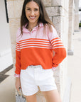 Woman wearing a pink and orange striped sweater with white shorts, standing against a stone wall.