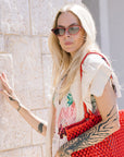 Woman wearing Solange Sunglasses Scarlet Chroma + Bordeaux holding a red woven bag against a stone wall