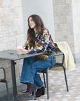 Woman sitting at an outdoor table with a cup, wearing a floral top and jeans.