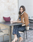 Woman sitting at an outdoor cafe table with a drink, wearing a brown jacket and floral pants. Lune Bag in Currant on table