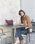 Woman sitting at an outdoor cafe table with a drink, wearing a brown sweater and floral set.