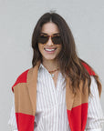Woman wearing a red and beige color-blocked cardigan over a striped shirt with Zander Hawksbill Sunglasses against a gray background