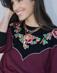 Woman wearing a burgundy sweater with floral embroidery and gold jewelry.