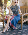 Woman sitting on a wooden chair wearing a gray t-shirt and blue patterned skirt.