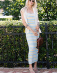 Woman in a striped dress and cardigan standing outdoors with greenery in the background
