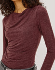 model wearing a long sleeve merlot top with a cowl neck and grey pants on a light grey background, close up