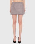A model from the waist down wearing a high-waisted, short A-line mini skirt in a fine red, black, and white gingham pattern, paired with black pointed-toe high heels, against a light gray studio background.
