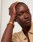 A close-up of a model wearing a beige with a gold bangle bracelet, against a plain light background.