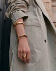 A close-up of a person wearing wide, polished gold bangle bracelets and a chunky gold ring, styled with a light beige trench coat.